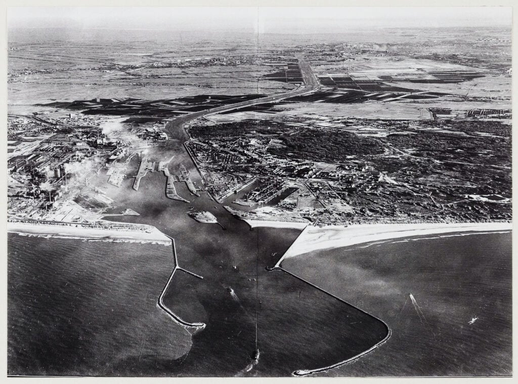 Luchtfoto van het Noordzeekanaalgebied, gezien in oostelijke richting. Op de voorgrond de pieren bij IJmuiden, op de achtegrond het IJsselmeer. Beeldcollectie van de gemeente Velsen